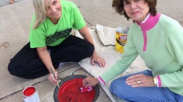 Two women paint a water meter cover red.
