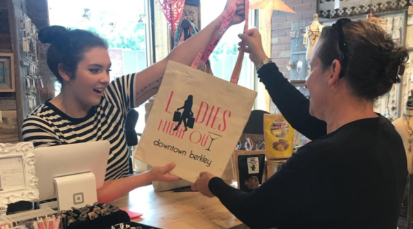 A young woman behind a desk at a small business hands a bag with words, "Ladies Night Out Downtown Berkley" to a customer.
