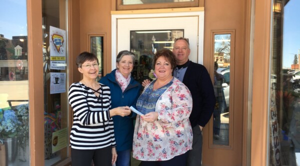Three women and one man stand in front of the front door of a small business holding a check.