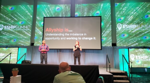 A man and woman give a presentation on a stage in front of big screen. On the screen, a slide shows the words, "Allyship is...Understanding the imbalance in opportunity and working to change it."