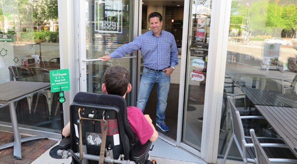 Man opens door of business for young man in wheelchair.