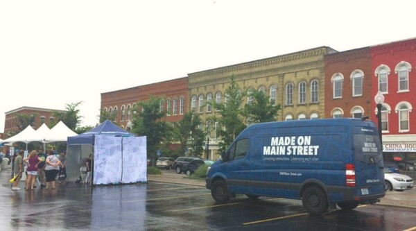 Blue van with words, "Made on Main Street" parked in front of historic buildings in a downtown street next to white pop-up tents and crowd of people.