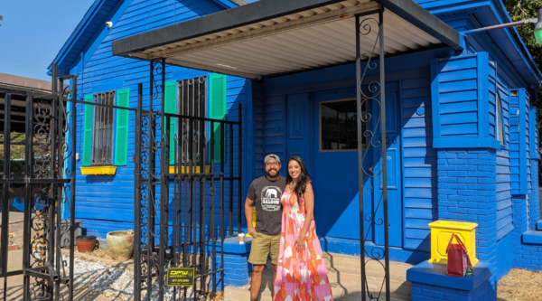 A man wearing a t-shirt and backwards cap and a woman wearing a pink dress smile in front of a blue house.