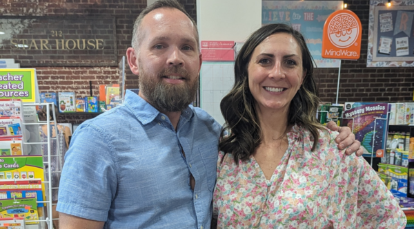 A man with a beard wearing a collared blue shirt and a woman with brown hair wearing a floral blouse smile inside a small business.