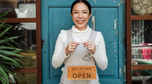 Woman holding sign called "Welcome We Are Open" in front of door