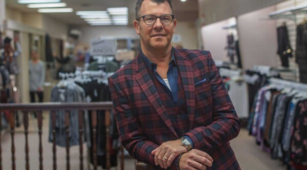 Un hombre con gafas y traje de chaqueta a cuadros se apoya en unas escaleras y sonríe, de pie en una tienda de ropa.