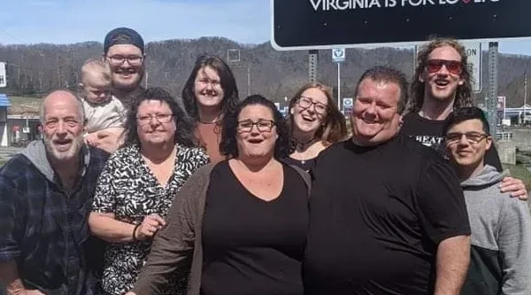 A group of people stand outside in front of a sign that says: "Welcome To Virginia: Virginia is For Lovers"