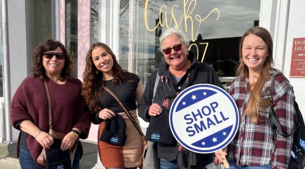 Cuatro mujeres posan con un cartel de Shop Small en una acera del centro de la ciudad