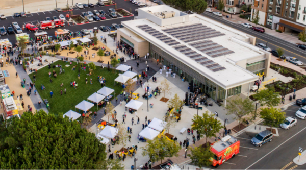People fill a large, open plaza dotted with booths and flanked with food trucks; new, 4-story mixed-use developments, trees, and gentle hills surround the plaza