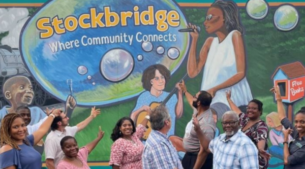A group of people pointing at a mural featuring bubbles, children playing, people playing music, and the text "stockbridge: where community connects"